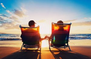 A photo showing a couple holding hands sitting in beach chairs near the ocean.