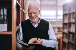 A photograph of a senior man smiling and reading a book in a library.