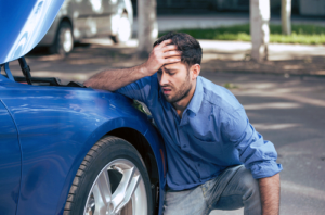 A photo showing a distressed man kneeling near his broken-down car.