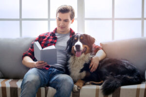 A photo of a young man attentively reading on a couch with his dog.