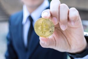 A photo showing a businessman in a dark suit holding a gold bitcoin.