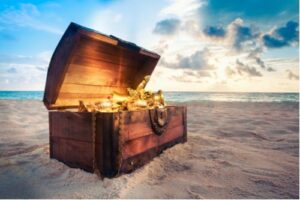 A photo showing a wooden treasure chest full of gold on a sandy beach near the ocean.