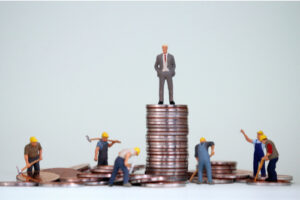 A conceptual image showing a businessman figure atop a tall stack of coins while blue-collar figures work among much shorter stacks around him.