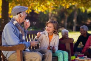 A photo of a grandfather sitting on a park bench with his granddaughter blowing bubbles.