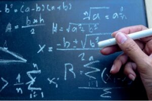 A photo showing a hand holding chalk and working on a complex mathematical equation on a blackboard.