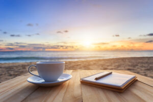 A photo showing a cup of coffee and a notebook resting atop a wooden table on the beach at sunrise.