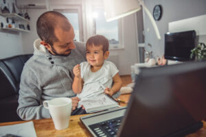 A photo showing a man working from home on his laptop while holding a child on his lap.