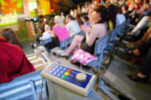 A photo showing a large studio audience with a voting device sitting on the knee of one audience member.