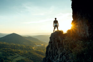 A photo of a man standing on the edge of a cliff looking out at a sunrise.