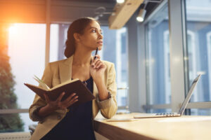 A photo of a young woman holding a notebook and looking out the window, thinking.