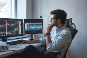 A man sits at a desk with multiple monitors showing stock charts.