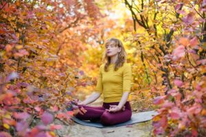 A photo showing a woman meditating outside in the woods with autumn leaves all around her.