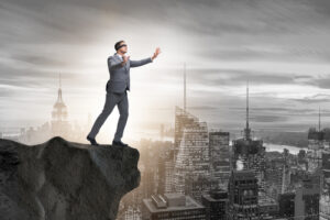 A businessman wearing a suit and blindfold stands near the edge of a cliff.