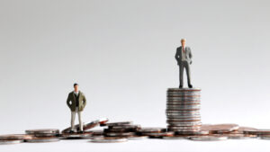 Two men stand on different-sized piles of coins, representing an imbalance of wealth.