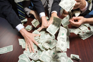 A photo showing a group of men in suits grabbing at a pile of money on a table.