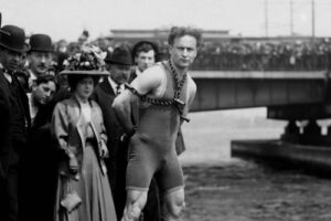 A black-and-white photo of Harry Houdini standing on the Harvard Bridge locked in chains and surrounded by onlookers.