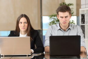 A photo of two employees working side by side and looking sideways at each other with envy.