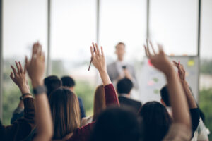 A photo showing a group of people in a business presentation with their hands raised.