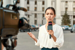 A photo showing a female reporter holding a microphone and filming a live report from a street.