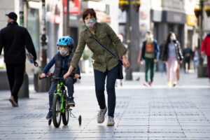 A photo showing a mother helping her son, both wearing masks, ride his bike down a commercial street.