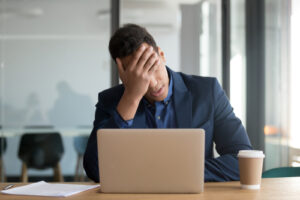 A businessman sits in front of his laptop with his palm to his forehead, looking stressed.