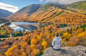 A photo showing a hiker sitting at the top of a bluff overlooking a lake and mountains.