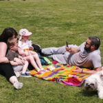 A photo of a father taking a picture of his wife and two young kids as they enjoy a picnic at the park.