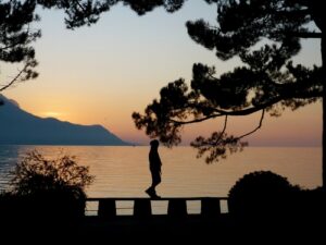 A photo of the silhouette of a woman walking peacefully along the side of a lake at sunset.