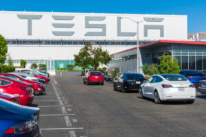 A photo of the parking lot of a Tesla manufacturing building in Silicon Valley with Tesla displayed on the exterior wall of the factory in large gray letters.