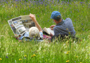 A photo of a couple resting in a meadow reading the morning newspaper together