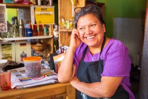 A photo of a woman who works at a coffee shop smiling in a black apron and purple shirt.