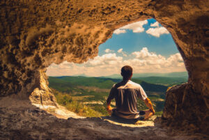 A photo of a man meditating in a cave while overlooking a mountain valley.