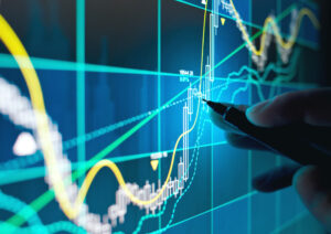 A close-up photo of an investment analyst’s hand tracking the technical movement of a stock chart on a computer screen with a pencil.