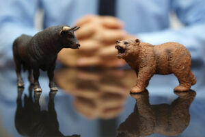 A close-up photo of two animal figurines - a bull and a bear - on a table. A businessperson has his hands folded together out of focus in the background.