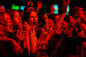 A photo of a group of young music fans standing in the front row of a concert with very expressive faces, yelling enthusiastically toward the performance.