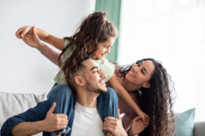 Parents enjoying quality time on the couch with their daughter as she smiles while on her father’s shoulders.