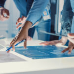A close-up of a small group of engineers gathered around a conference table analyzing blueprints for a new design.