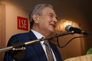 George Soros standing at the podium speaking into a microphone at the London School of Economics Alumni Dinner in Malaysia.