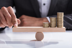 A close-up of a wooden seesaw, with three stacks of coins on one side and a businessperson pressing his finger on the other to keep it level.