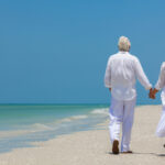A couple wearing all white walks hand in hand down the beach on a warm, sunny day.