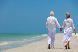 A couple wearing all white walks hand in hand down the beach on a warm, sunny day.