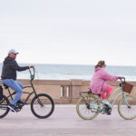 A photo of a man in a gray sweater and a woman in a pink coat riding bikes along the boardwalk on a chilly afternoon.