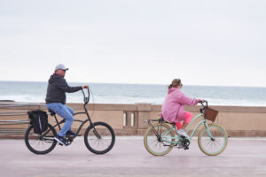 A photo of a man in a gray sweater and a woman in a pink coat riding bikes along the boardwalk on a chilly afternoon.