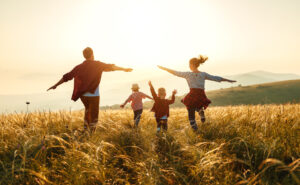 Parents run with their two children through a field of long grass at sunset.