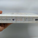 A hand holding Malcolm Gladwell’s book Outliers.
