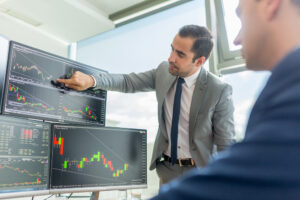 Two stockbrokers looking at stock indexes on multiple computer screens in a trader’s office.