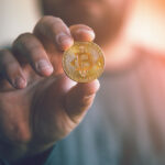A man holding up a gold coin with the Bitcoin symbol on the face.