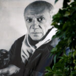 A black-and-white portrait of Pablo Picasso hung up behind a green tree.