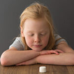 A child sits at a table across from a single marshmallow.