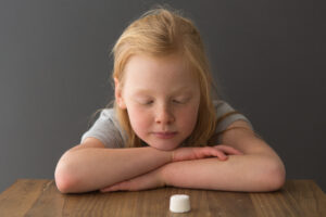 A child sits at a table across from a single marshmallow.